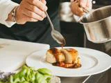 En kok hælder sauce over en fyldt kyllingeroulade på en hvid tallerken, pyntet med grøntsager, i et professionelt køkken.