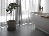 En potteplante står på et flisegulv ved siden af et moderne hvidt badekar med gennemsigtige gardiner i baggrunden.
