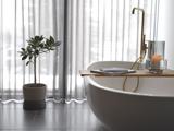 Moderne badeværelse med hvidt badekar, vandhane i guld, træbakke med bog og stearinlys, potteplante og gennemsigtige gardiner, der lukker naturligt lys ind.
