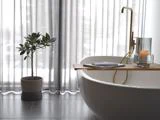 Moderne badeværelse med hvidt badekar, vandhane i guld, træbakke med bog og stearinlys, potteplante og gennemsigtige gardiner, der lukker naturligt lys ind.