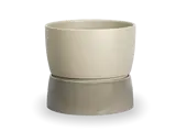 En enkel, rund, beige urtepotte, der står oven på en matchende, lidt bredere underkop på en ensfarvet baggrund.