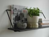 En sort væghylde rummer en kogebog, en potteplante, en rund sten og stablede bøger mod en hvid væg.