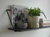 En sort væghylde rummer en kogebog, en potteplante, en rund sten og stablede bøger mod en hvid væg.
