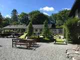 Et solrigt udendørsområde med bænke, liggestole, træer og en minigolfbane ved siden af en hvid bygning omgivet af grønt.