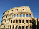 Colosseum i Rom, Italien, under en klar blå himmel, hvor sollyset oplyser de gamle stenbuer og et par høje træer i forgrunden.