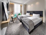 Moderne hotelværelse med to enkeltsenge, grå hovedgærder, hvidt sengetøj, mønstrede plaider, et skrivebord med to stole, et vindue med gardiner og indrammet kunst på væggen.