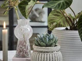 Et gråt stearinlys, en vase med blomster og grønne potteplanter står på et bord og skaber et hyggeligt, naturligt indretningsarrangement.