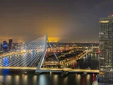 Et natligt bybillede med Erasmus-broen i Rotterdam, oplyste bygninger og refleksioner i floden under en glødende himmel.