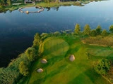 Luftfoto af en golfbanegreen med tre sandbunkere nær en rolig sø, omgivet af træer og frodigt græs.