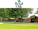 Moderne hytter med grønne tage er placeret blandt frodige træer og græs under et hvidt banner med Royal Oak-logoet og en grafik af egeblade.