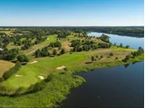 Luftfoto af en grøn golfbane med sandbunkers ved siden af en rolig flod, omgivet af træer og åbne marker under en klar blå himmel.