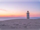 Et ensomt fyrtårn står på en sandklit ved solnedgang med udsigt over havet.