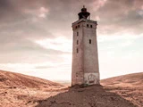 Et højt, forvitret fyrtårn står alene på sandet, gold jord under en overskyet himmel med blødt sollys.