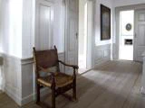 En træstol med et sæde af brunt læder står i en lys entre med trægulv, hvide paneler på væggene og indrammede portrætter, der hænger på væggene.