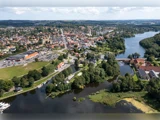 Luftfoto af en lille by med bygninger med røde tage, grønne træer, parkeringspladser og flere søer og floder, der snor sig gennem landskabet under en delvist overskyet himmel.