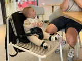 En smilende baby i beige tøj sidder sikkert i en sele på en stol og kigger op på en voksen, der sidder ved siden af dem ved et bord i et lyst indendørs miljø.
