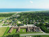 Luftfoto af en kystby med huse, grønne træer og marker, der fører til en sandstrand og blåt hav under en klar himmel.