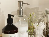 En sæbedispenser, en lotionflaske, en lille vase med kamilleblomster, glaskrukker og vatpinde arrangeret pænt på en bakke.