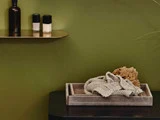 En beige bakke med en strikket klud og en naturlig svamp står på en mørk overflade; ovenover holder en guldhylde to neglelakflasker og en brun flaske mod en olivengrøn væg.