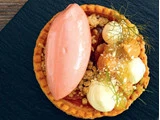 En tærte toppet med en kugle lyserød mousse, hvide flødeboller, karamelliseret frugt, krummer og frisk urtegarniture på en mørk skiferoverflade.