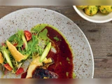 Nærbillede af en gourmetret på en plettet tallerken med salat, sauce og pynt, med en lille skål kartofler i baggrunden på et træbord.