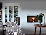 En hyggelig restaurant med vinglas på bordene, en tændt pejs, hylder med flasker og et rundt bord med en vase med blomster og et stearinlys.