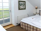 Et hyggeligt soveværelse med en pænt redt seng, et natbord med en lampe, et indrammet botanisk tryk på væggen og en glasdør, der fører ud til en balkon med udsigt over grønne marker.