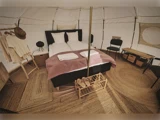 Et hyggeligt glampingtelt med dobbeltseng, lyserødt tæppe, håndklæder, stole, træopbevaring, vævede tæpper og pænt arrangeret tøj og tøfler.