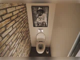 Et toilet med åbent låg er placeret under et indrammet sort-hvidt foto af en mand med hat, der er hængt op på en almindelig væg ved siden af fritlagt murværk. En toiletbørste står ved siden af toilett