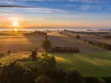 Luftfoto af et landskab ved solopgang med en traditionel vindmølle, marker, spredte træer og sollys, der kaster lange skygger over landskabet.