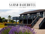 Et mørkt strandhotel i træ med et øvre dæk og trapper, omgivet af blomstrende lilla lavendler under en overskyet himmel; på skiltet står der Samsø Badehotel Ilse Made.