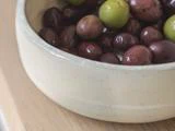 Et nærbillede af en beige keramikskål fyldt med forskellige grønne og lilla oliven, placeret på en lys træoverflade.