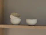 Tre små hvide keramikskåle på en træhylde; to er stablet med den øverste på skrå, og en skål står for sig selv.