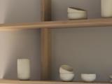 Minimalistiske træhylder med enkle, håndlavede hvide keramiske kopper og skåle, med dæmpet belysning og en neutral væg i baggrunden.