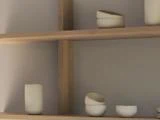 Minimalistiske træhylder med enkle, håndlavede hvide keramiske kopper og skåle, med dæmpet belysning og en neutral væg i baggrunden.