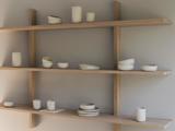 Tre træhylder viser minimalistiske hvide keramikvaser, skåle og kopper i forskellige former og størrelser mod en lysegrå væg.