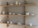 Tre træhylder viser minimalistiske hvide keramikvaser, skåle og kopper i forskellige former og størrelser mod en lysegrå væg.
