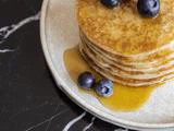 En stak pandekager toppet med blåbær og sirup, serveret på en hvid tallerken på en sort marmoroverflade.