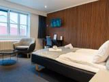 Moderne hotelværelse med dobbeltseng, lænestol, skrivebord, tv monteret på en accentvæg af træ, rundt glasbord og stort vindue, der lukker naturligt lys ind.