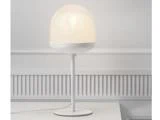 En moderne hvid bordlampe med en rund, matteret glasskærm står på en hvid flade mod en hvid panelvæg.