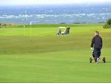 En mand skubber en golfvogn på en grøn golfbane nær havet, med to andre mennesker og en golfvogn i det fjerne.