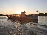 En hvid båd ved navn Viking sejler over roligt vand ved solnedgang, mens solen skinner bag den, og træerne er synlige i baggrunden.