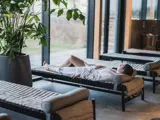 En person i hvid badekåbe slapper af på en liggestol i en moderne indendørs spa med store vinduer og grønne planter i nærheden.
