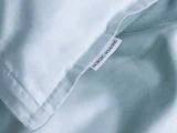 Nærbillede af et lyseblåt stof med et synligt NORDIC WEAVING-mærke syet ind i sømmen, hvilket tyder på sengetøj, f.eks. et pudebetræk eller et dynebetræk.