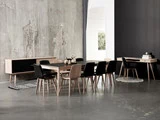 Minimalistisk spisestue med træbord, otte sorte stole, skænk, skrivebord og betonvægge; blødt lys kommer ind fra et tilstødende rum.