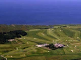 Luftfoto af en golfbane med sandbunkers, grønne fairways, et klubhus og en parkeringsplads, der ligger ved en kystlinje med blåt hav bagved.