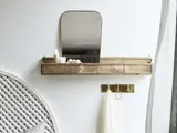 En guldhylde med spejl, toiletsager og skaller over en guldknagerække med en hvid klud, monteret på en hvid væg ved siden af en seng med en hvid vævet hovedgærde.