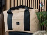 En beige duffelbag med sorte håndtag står på en mørk flade med en logopatch på forsiden. I baggrunden er der trælister og grønne planter, og et Atelier Sheworks-skilt er synligt.
