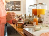 En person i lyserødt ærme øser toppings på yoghurt ved en morgenmadsbuffet med krukker med juice, morgenmadsprodukter og krydderier på en trædisk.