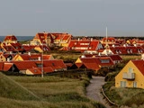 En kystby med gule huse og røde tage omgivet af græsklædte klitter under en overskyet himmel.